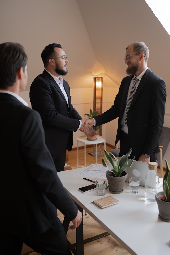 Businessmen in formal attire shaking hands during a meeting in a modern office setting.
